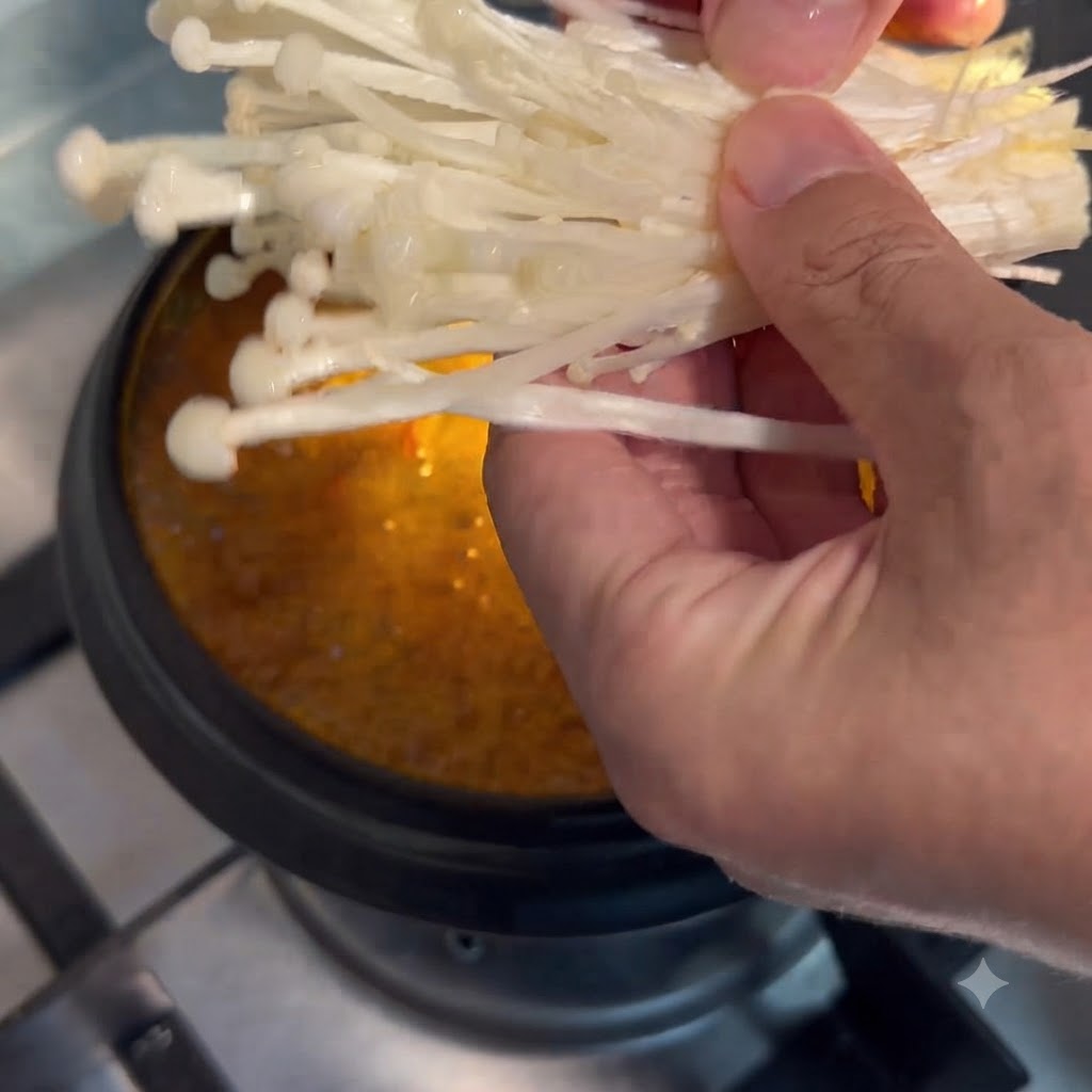 Enoki mushrooms being held by hand and lowered into the simmering pot of soup.