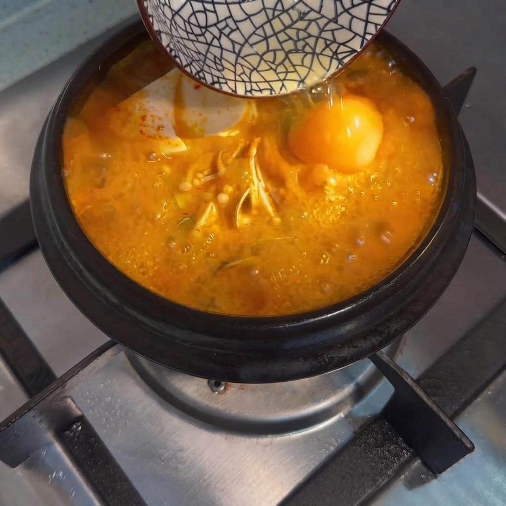 A raw egg being poured from a small bowl into the bubbling Korean tofu stew.