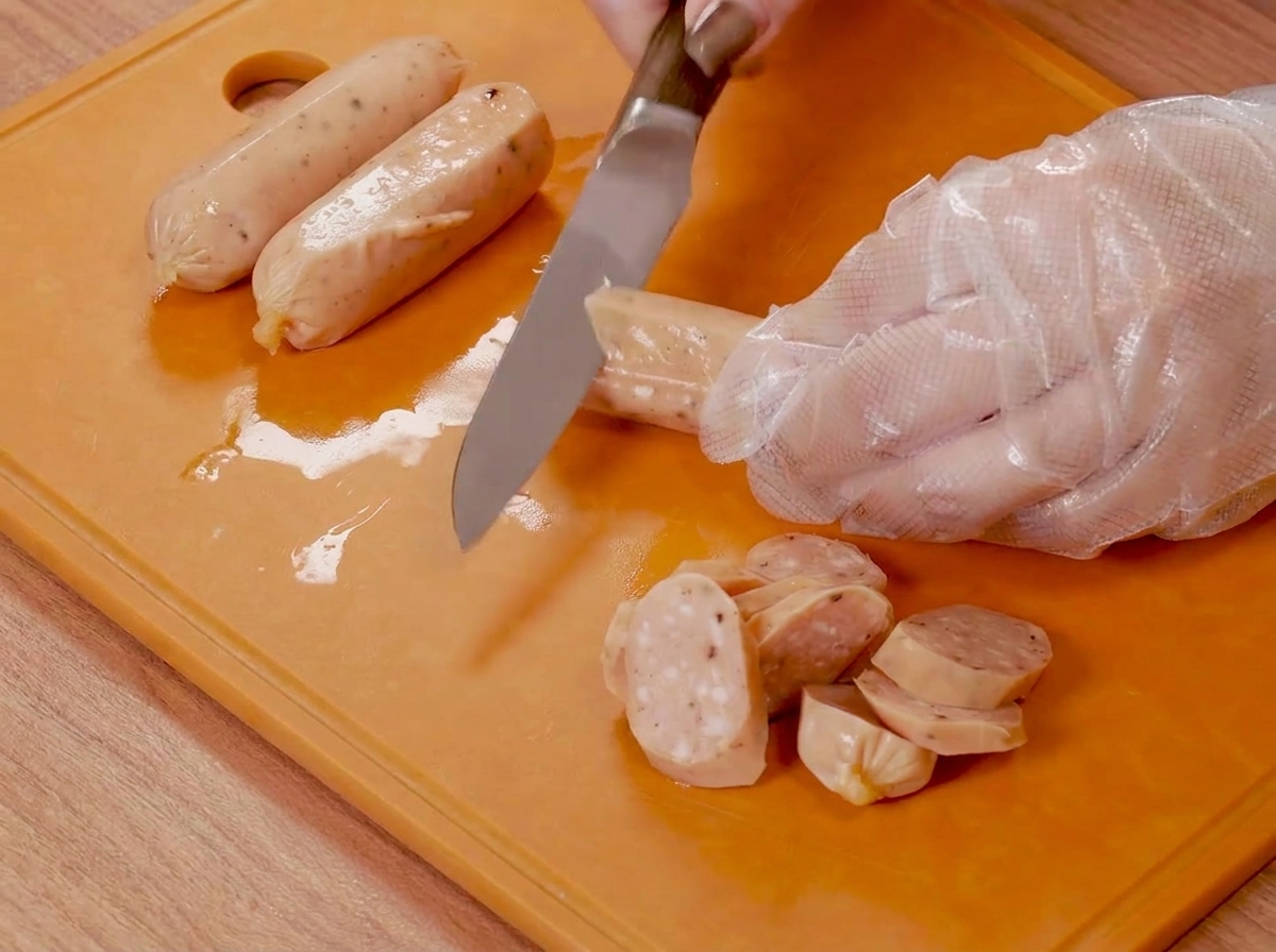 Hands wearing plastic gloves using a knife to score small sausages on an orange cutting board.