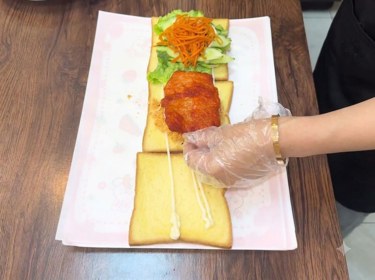 A gloved hand placing a warm fried chicken cutlet onto the layered toast assembly.