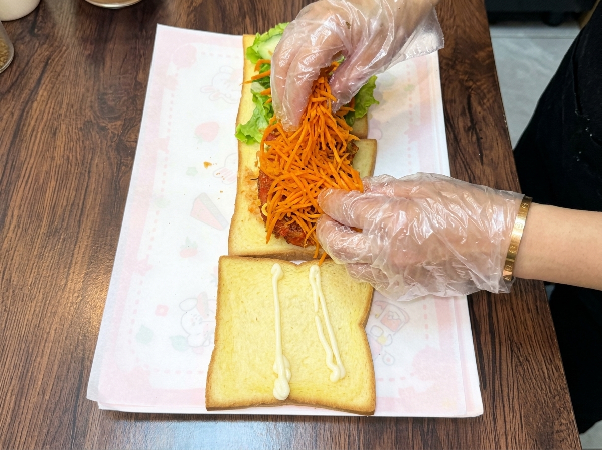 Gloved hands piling a generous mound of bright orange, shredded stir-fried carrots on top of the stacked chicken cutlet.
