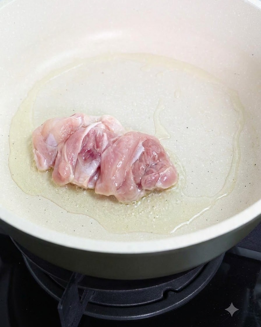A raw chicken thigh frying skin-side down in an oiled pan.