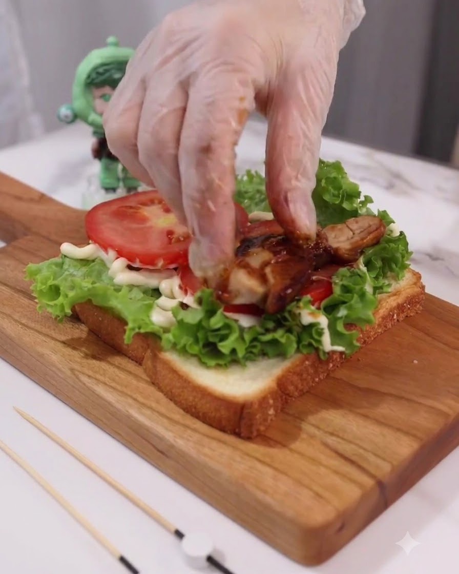 Gloved hand placing cooked teriyaki chicken slices onto the tomato and lettuce layers.