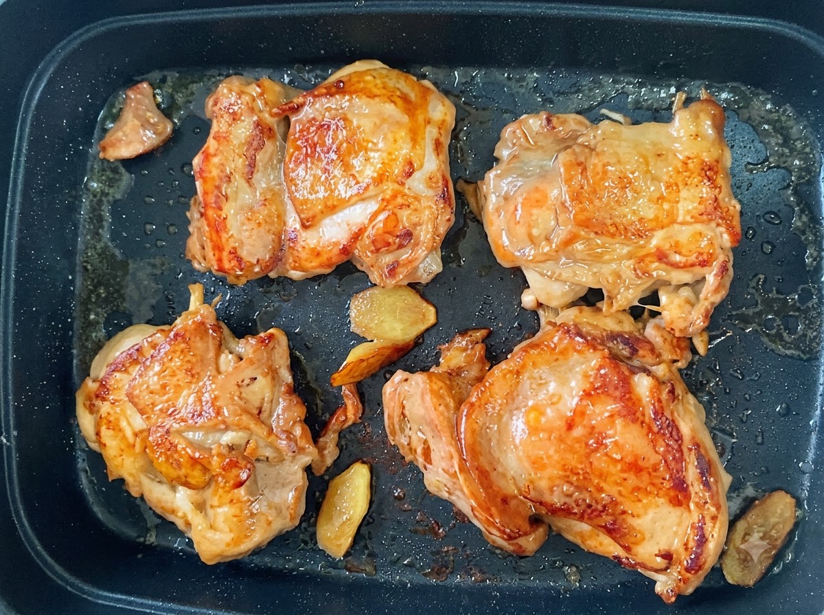 Four chicken thighs frying in a pan with golden brown crispy skin and ginger slices.