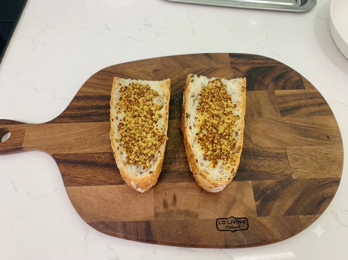 Two slices of rustic bread placed on a wooden cutting board, both evenly spread with seeded whole grain mustard.