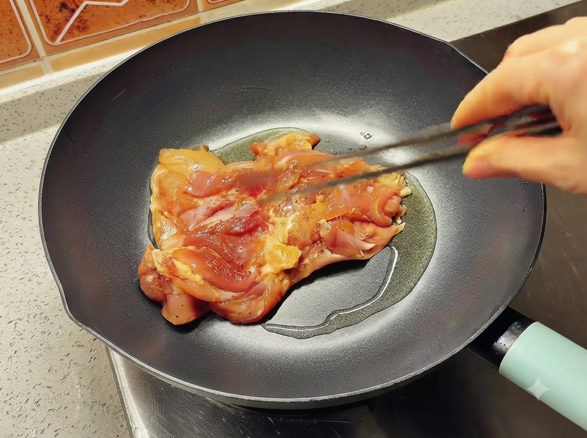 A single marinated chicken thigh piece being placed into a hot oiled skillet using tongs.