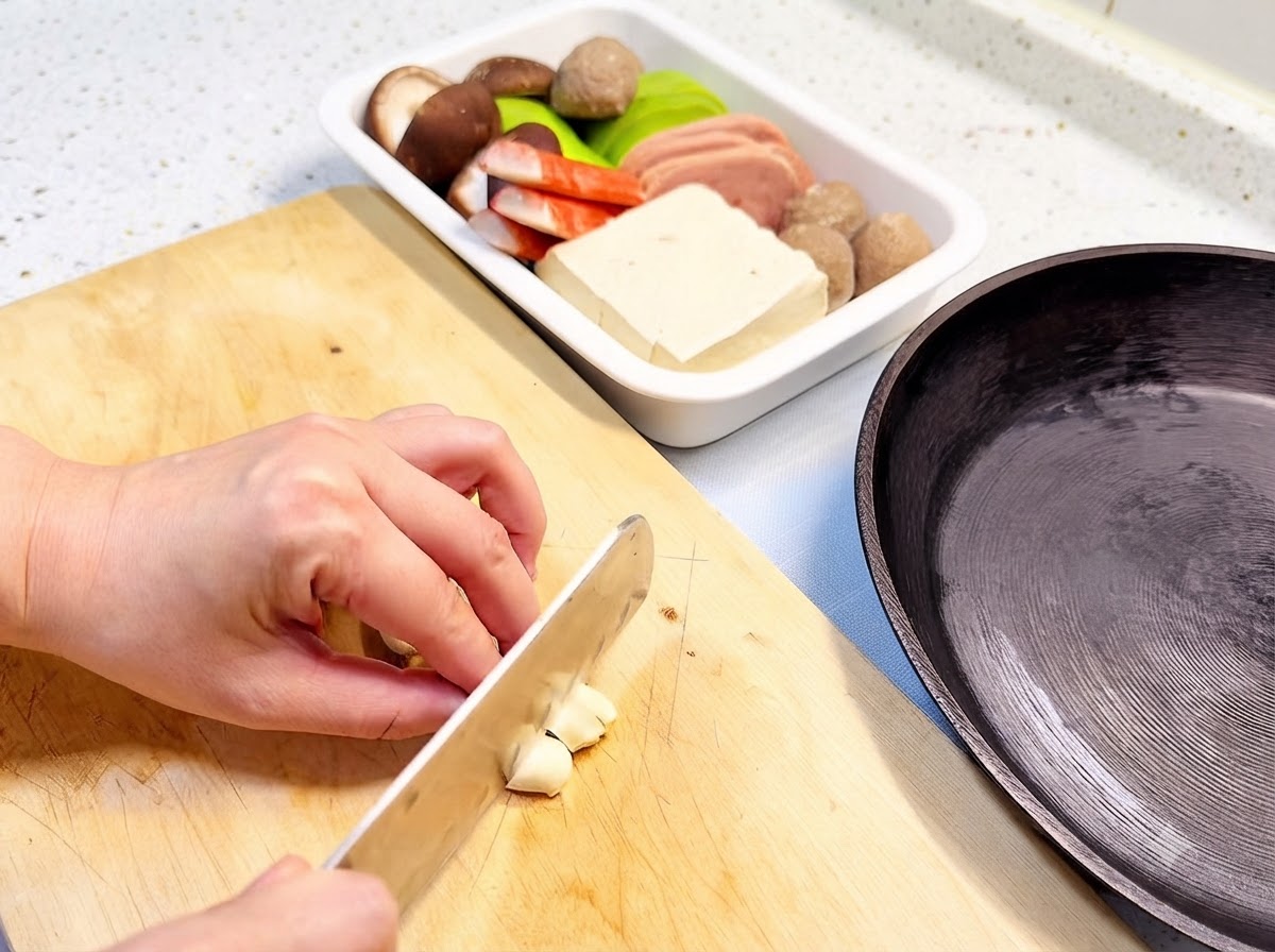 Garlic cloves being sliced on a wooden cutting board next to a frying pan and an ingredient tray.