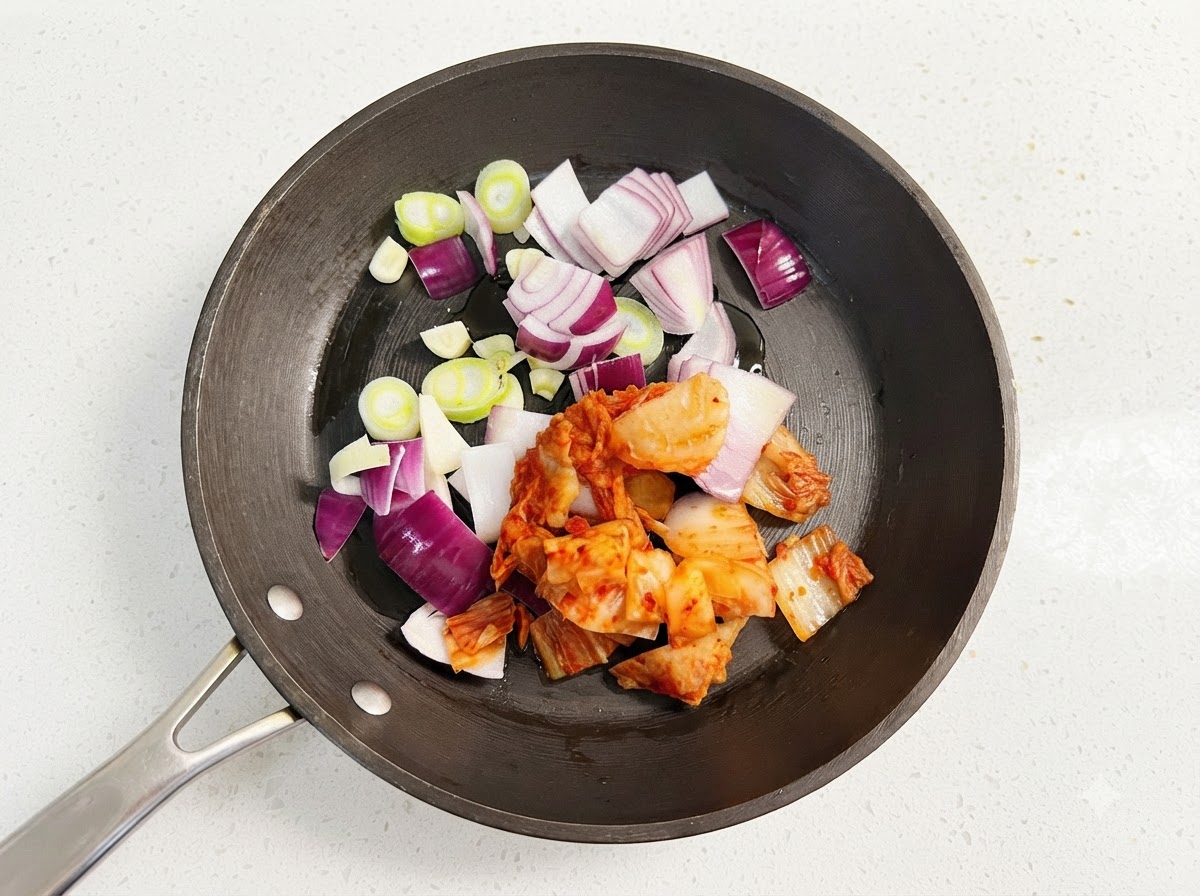 Slices of purple onion, green scallion rounds, and chopped red kimchi sizzling in a black skillet with oil.