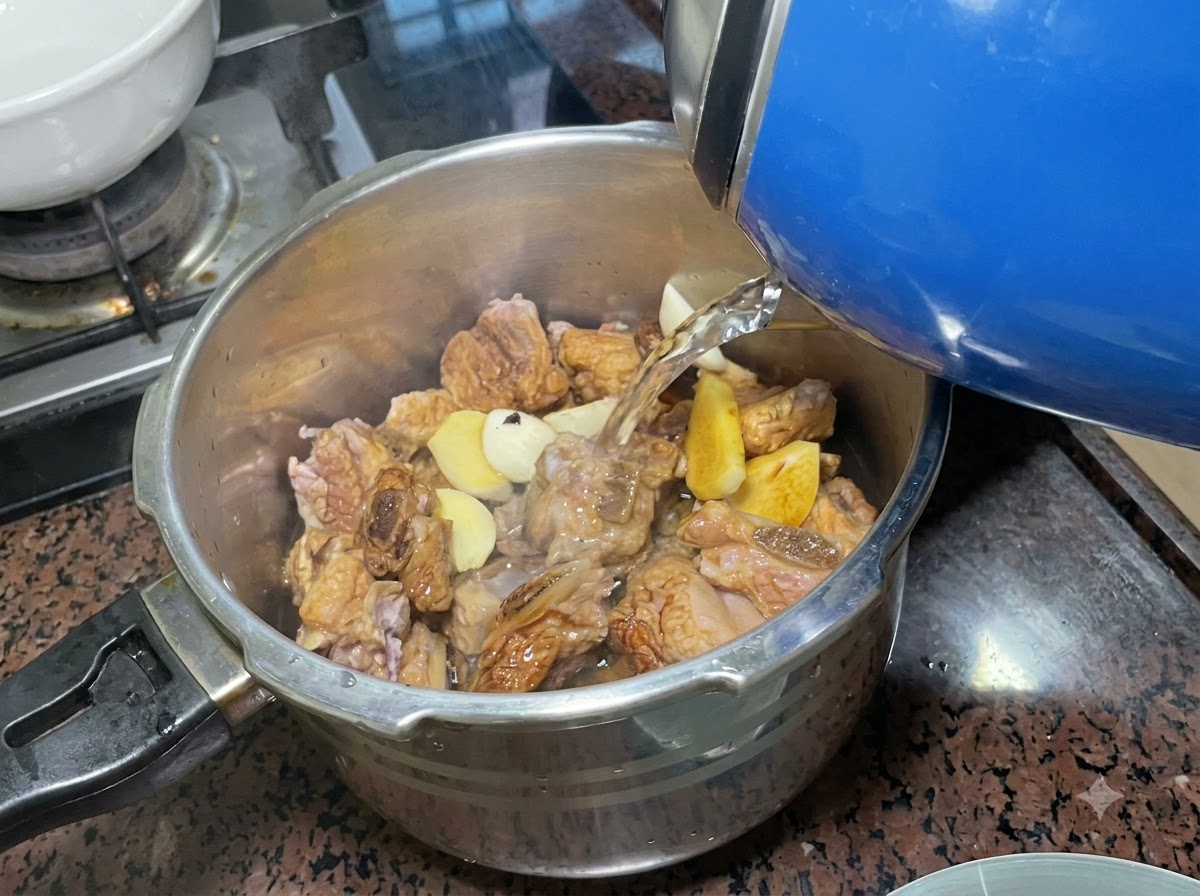 Water being poured from a blue electric kettle into a stainless steel pressure cooker filled with seasoned ribs and aromatics.