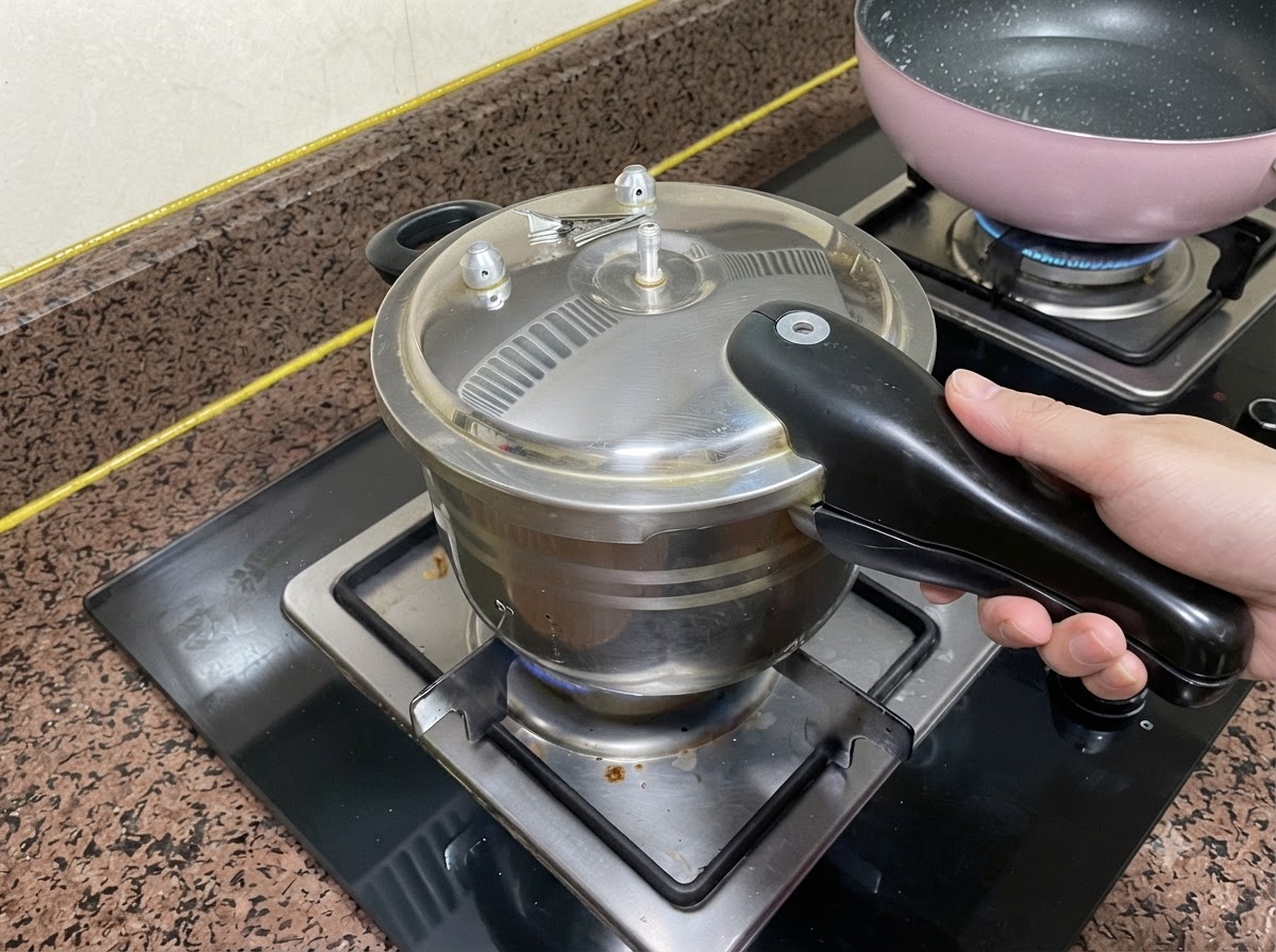 A pressure cooker lid closed on a stovetop burner with a hand gripping the handle.
