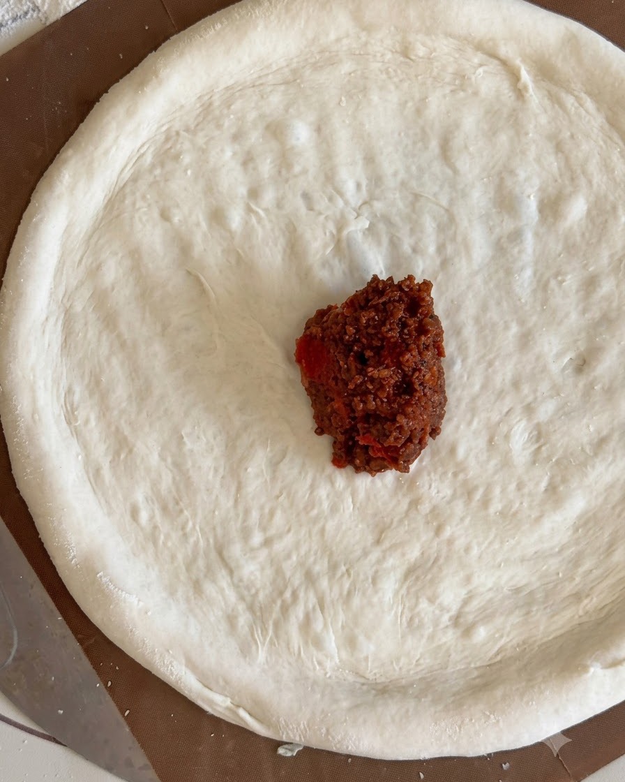 A small mound of dark red meat sauce sitting in the center of a flat pizza dough circle.