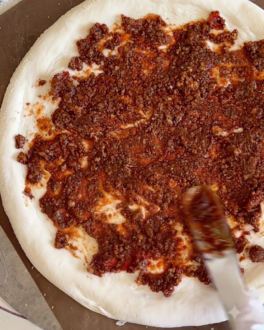 Cooked beef cubes being scattered over the red meat sauce spread evenly across the white pizza dough base.