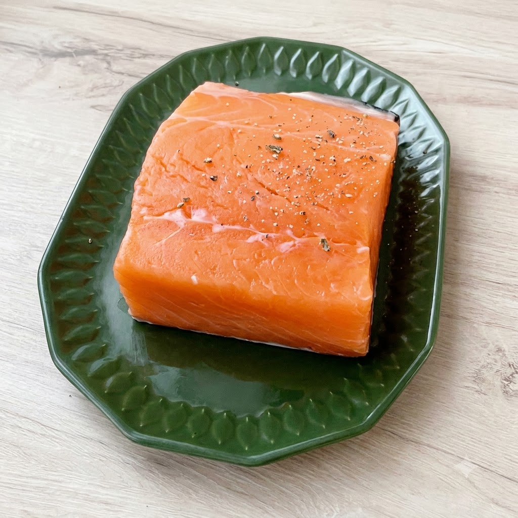 A thick piece of fresh salmon sitting on a green plate, generously dusted with coarse sea salt and cracked black pepper.