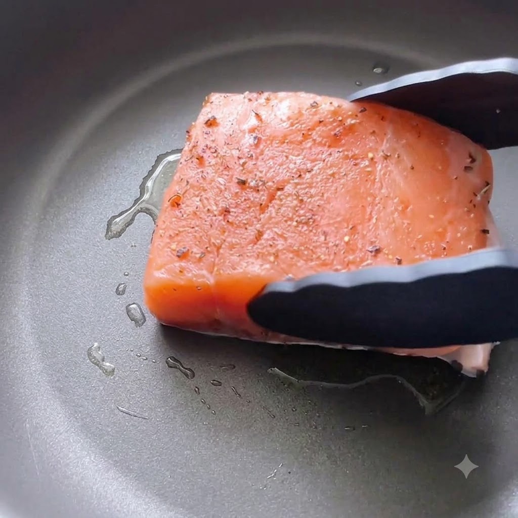 A seasoned salmon fillet being lowered skin-side down into an oiled skillet using black kitchen tongs.