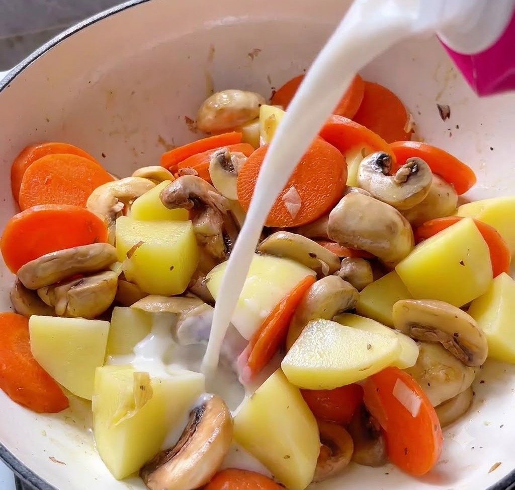 Liquid being poured from a pink carton into a cooking pot filled with potato cubes, carrot slices, and mushrooms.