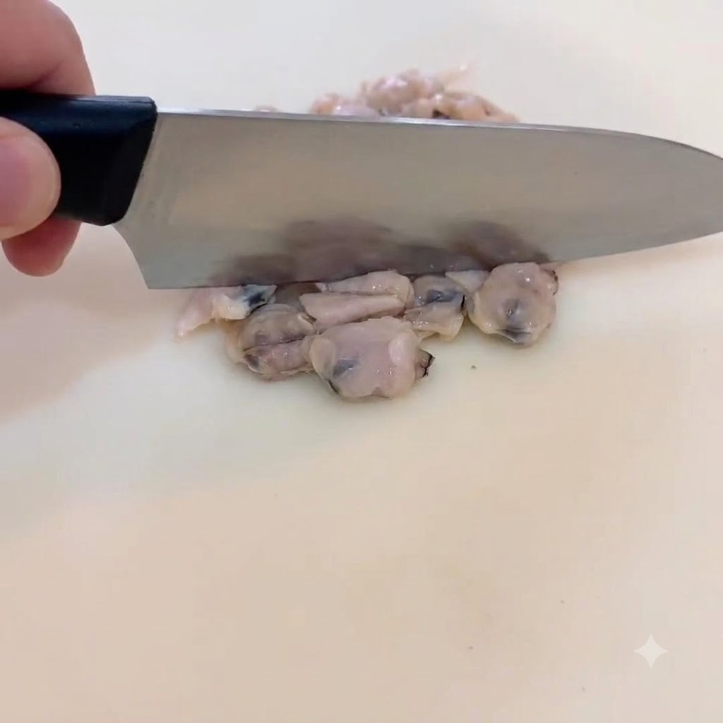 A close-up of a hand holding a chefs knife, gently chopping cooked clam meat into small, bite-sized pieces on a white cutting board.