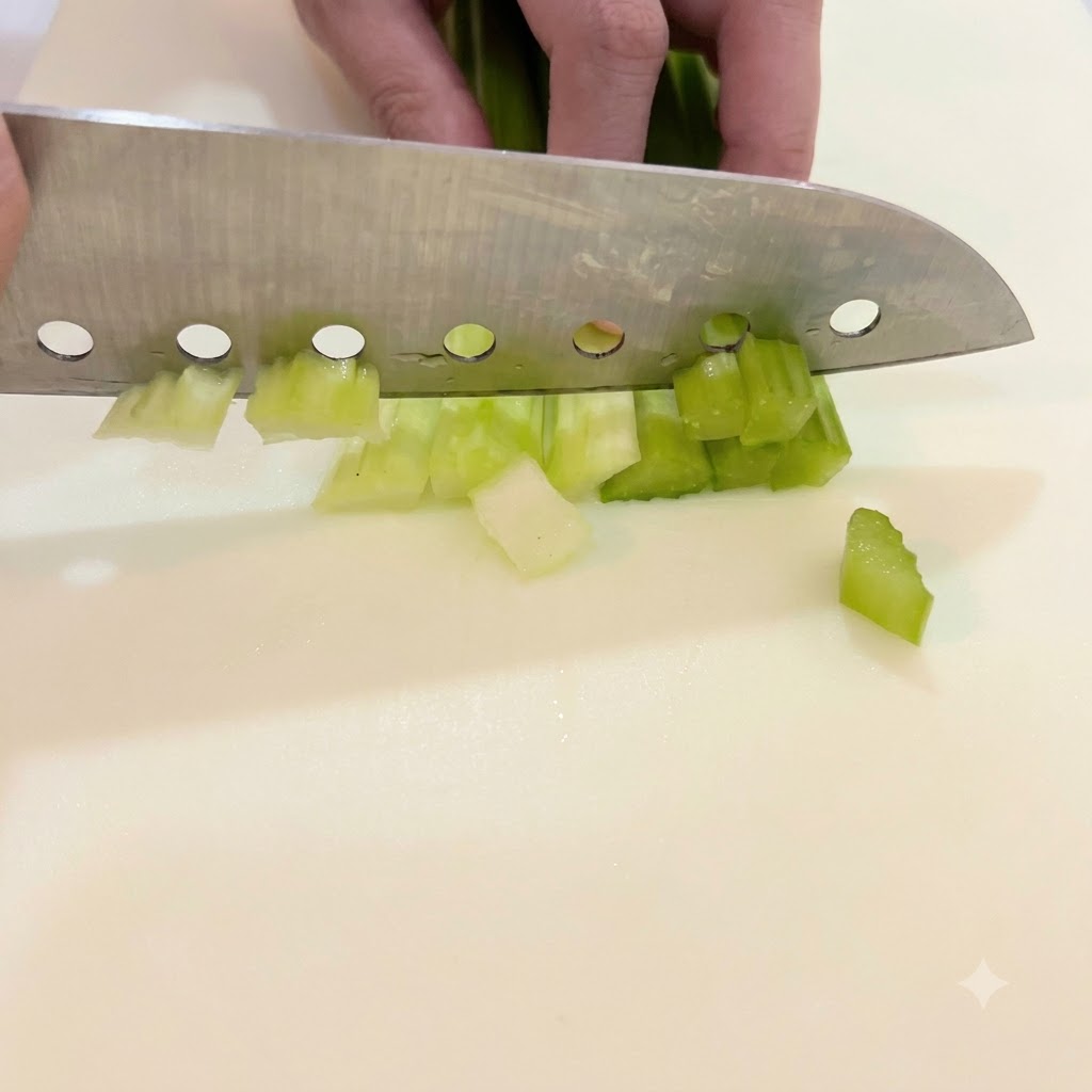 A hand guiding a large knife to chop bright green celery stalks into small, even chunks on a white cutting board.