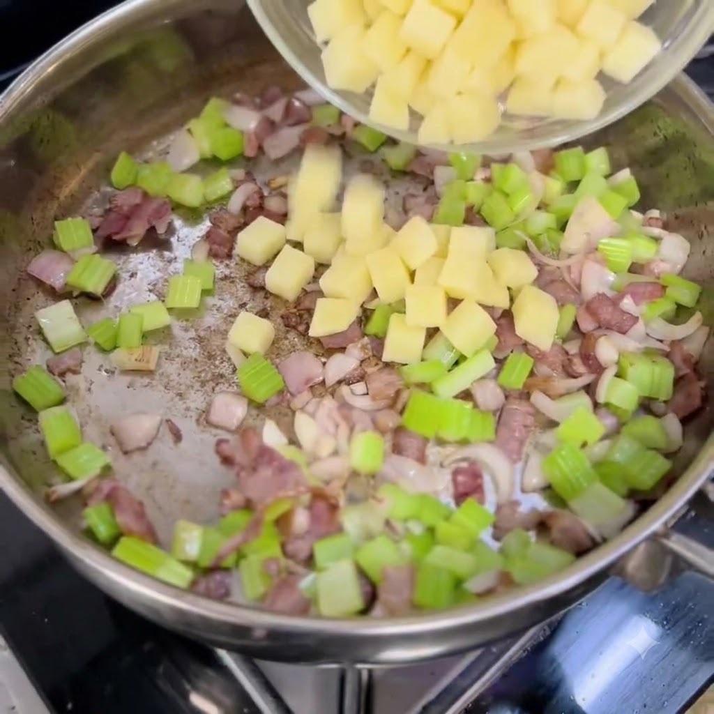 Diced potatoes being poured from a clear bowl into a pan containing chopped onions, celery, and bacon.