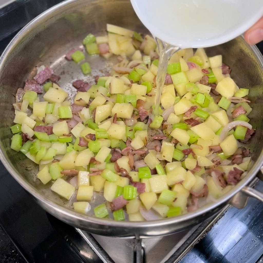 Clear clam broth being poured from a white bowl into a pan of sautéed vegetables and bacon.