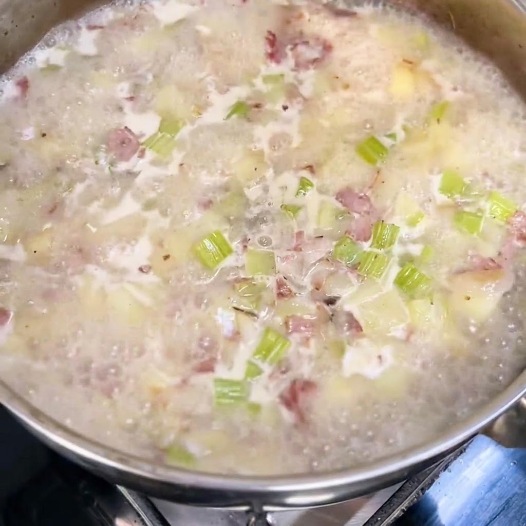 Milk and heavy cream being poured into a simmering pot of soup with visible chunks of potatoes, celery, and bacon.