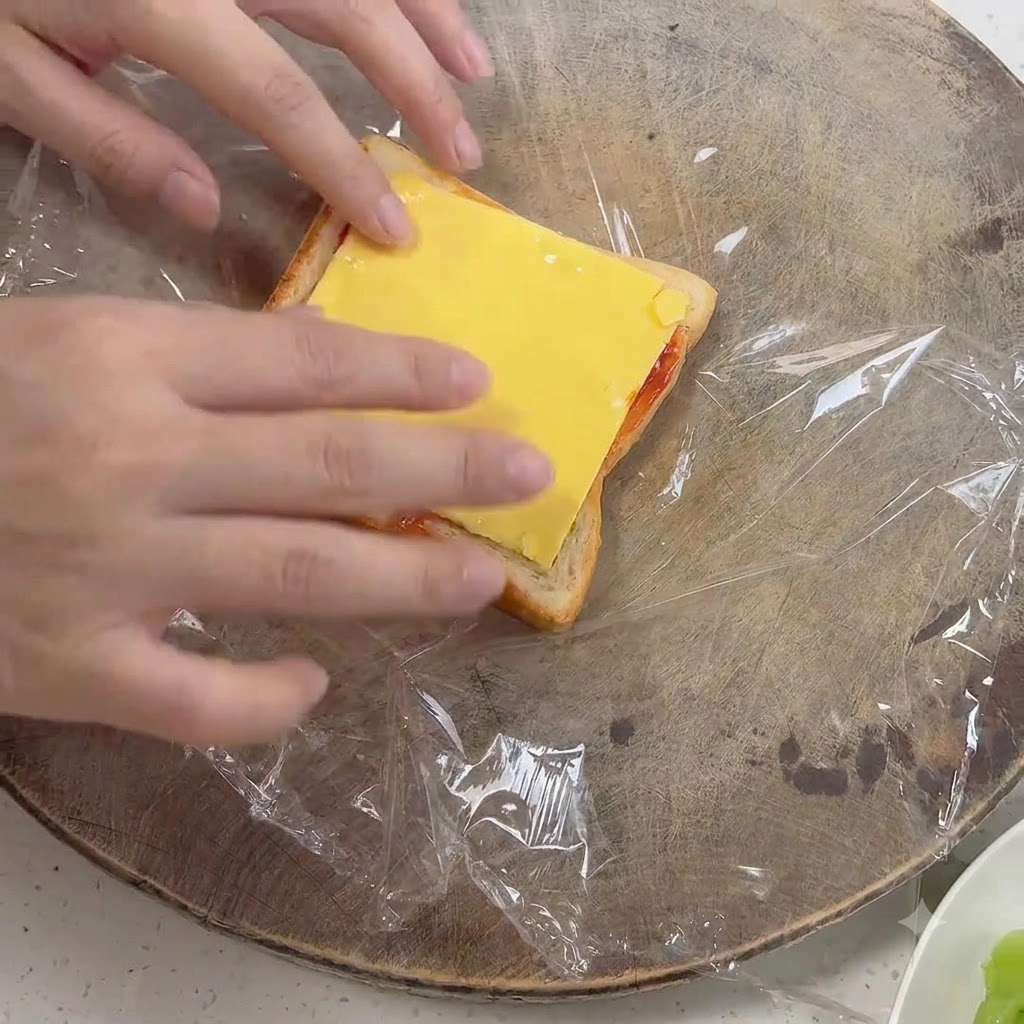 Hands placing a yellow square cheese slice onto a piece of toast covered with sauce.