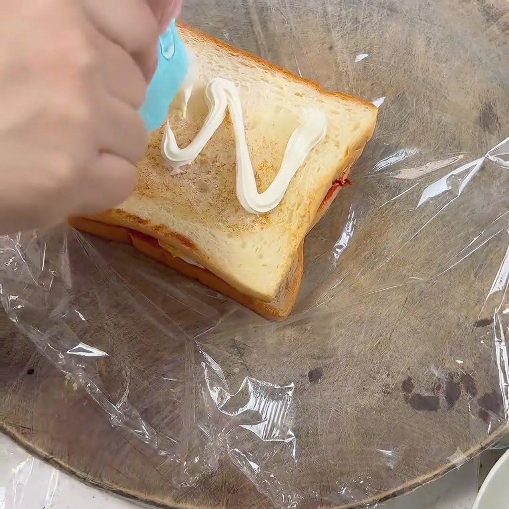 Squeezing mayonnaise in a zigzag pattern onto a slice of toasted bread.