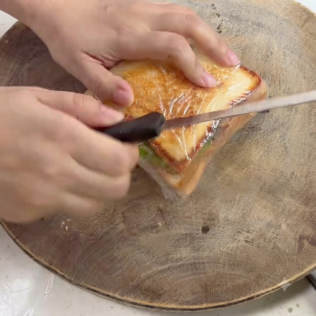 A kitchen knife slicing diagonally through a plastic-wrapped sandwich on a wooden board.