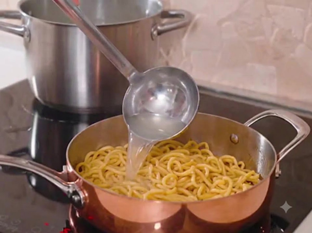 A metal ladle pouring starchy pasta water into a shiny copper pan with cooked spaghetti.