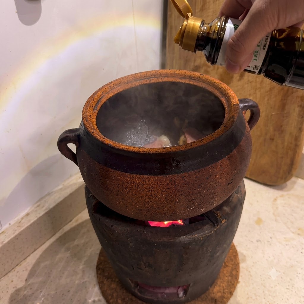 Soy sauce being poured from a bottle into a clay pot sitting on a burning charcoal stove.
