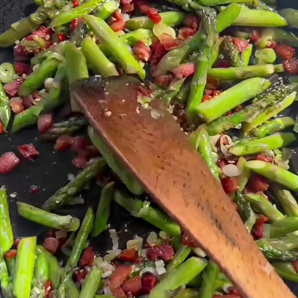 Clear liquid stock being poured from a container into a sizzling pan filled with sliced green asparagus and bacon pieces.