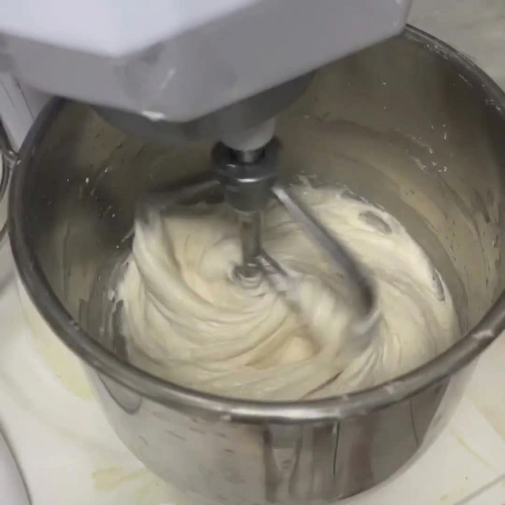 White dough combining with liquid inside a large stainless steel stand mixer bowl using a dough hook.