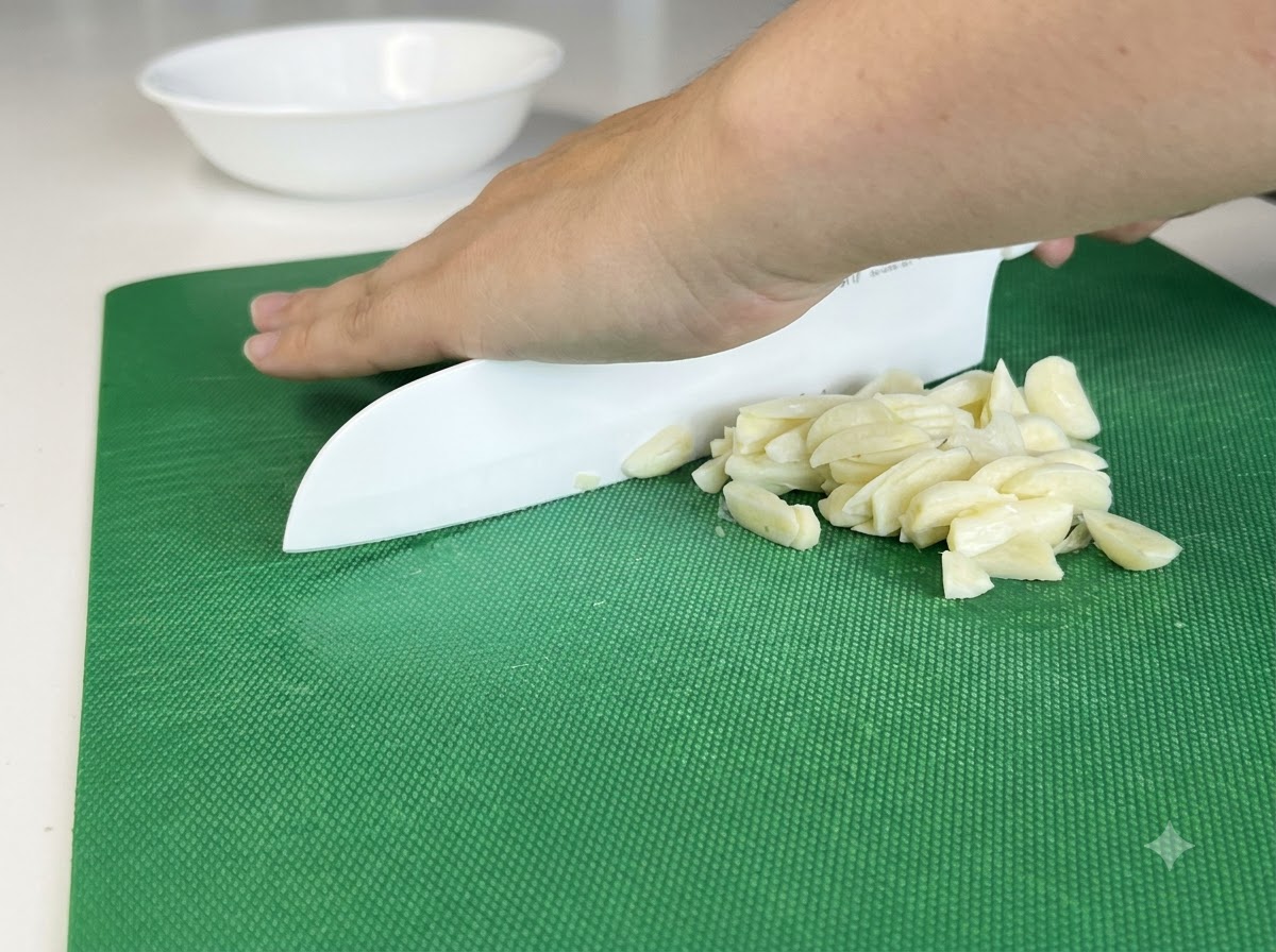 Garlic cloves being finely chopped on a cutting board using a white knife.