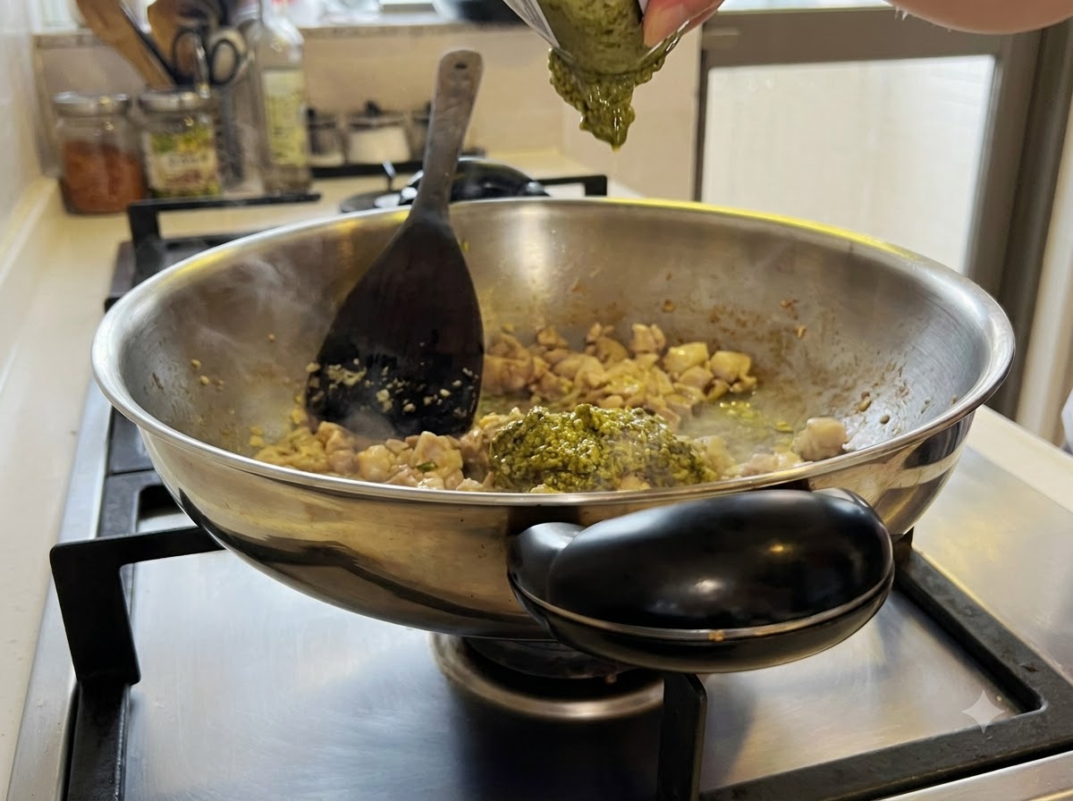 Green pesto sauce being mixed into the chicken and garlic mixture inside the wok.