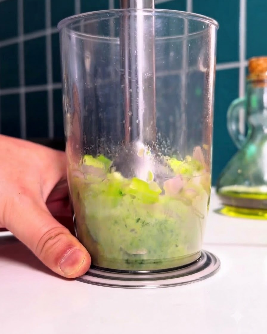 An immersion blender pureeing blanched green vegetables and spinach inside a tall clear cup.