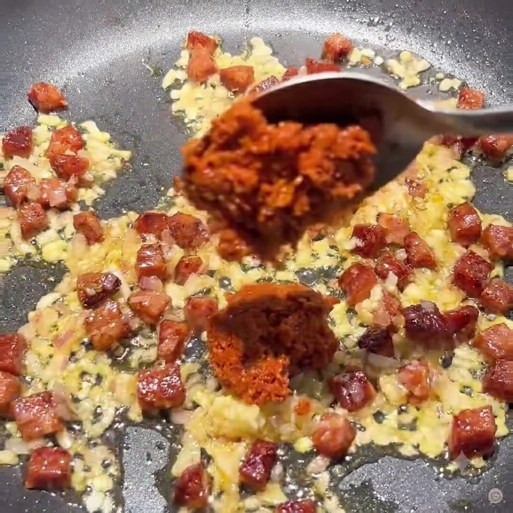 Spoon adding thick sun-dried tomato paste and tomato paste into the pan with the chorizo and onion mixture.
