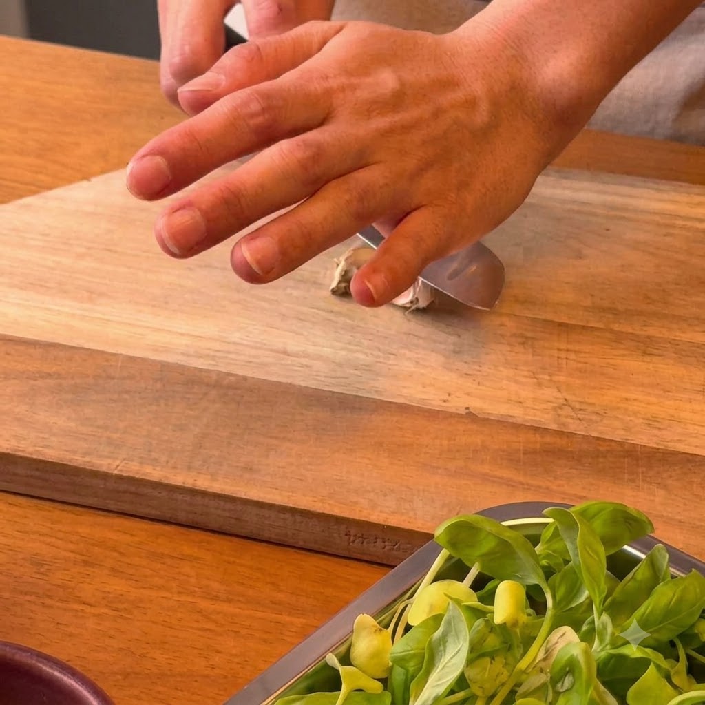 A hand pressing the flat side of a chefs knife down onto a garlic clove on a wooden cutting board.