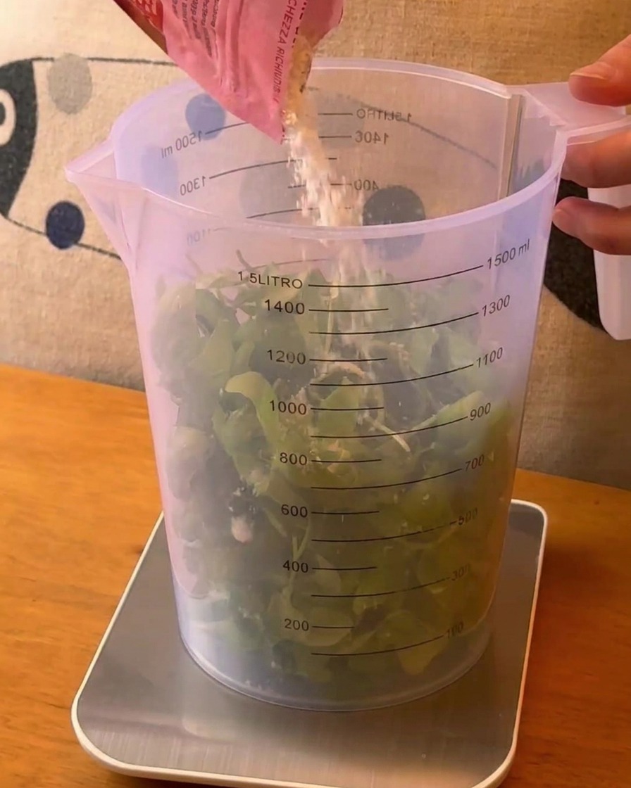 Grated cheese being poured from a pink pouch into a large measuring cup filled with fresh green basil leaves.