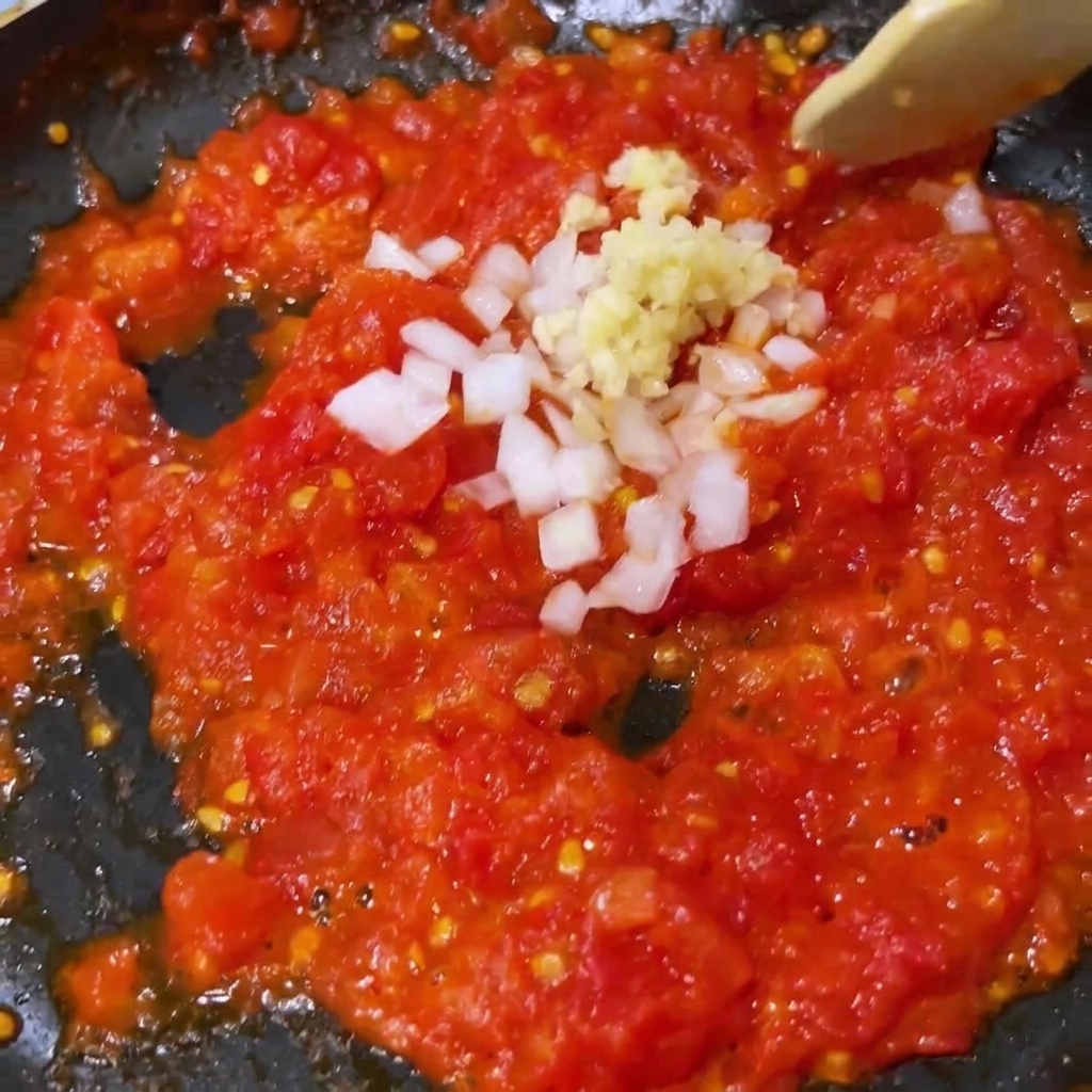 Minced garlic and white onion being mixed into softened crushed tomatoes in a pan.