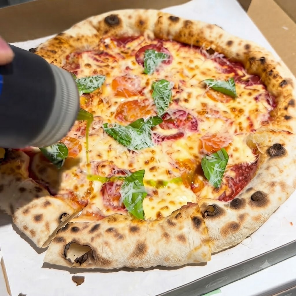 A bottle pouring a stream of extra virgin olive oil onto a freshly baked pizza topped with basil and cheese inside a box.