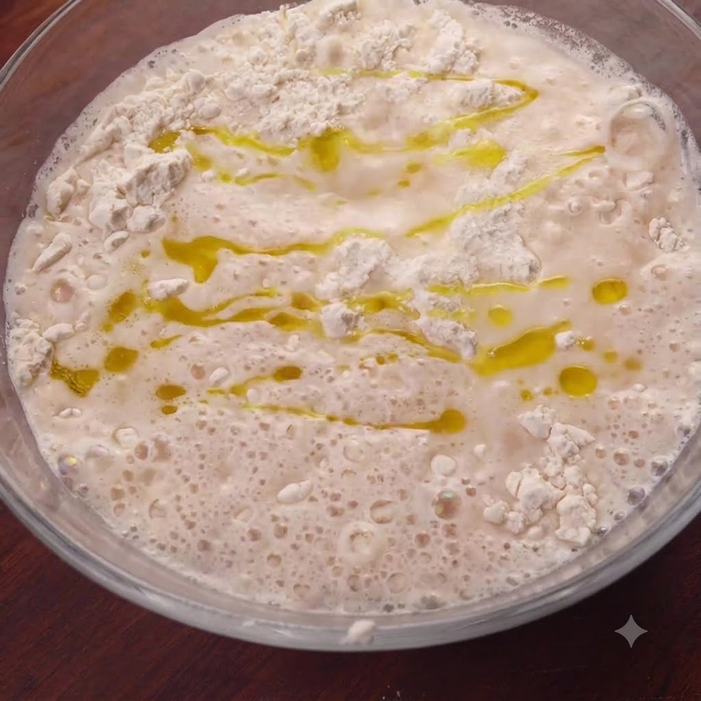 Golden olive oil drizzled across the surface of the wet flour mixture inside a glass bowl.