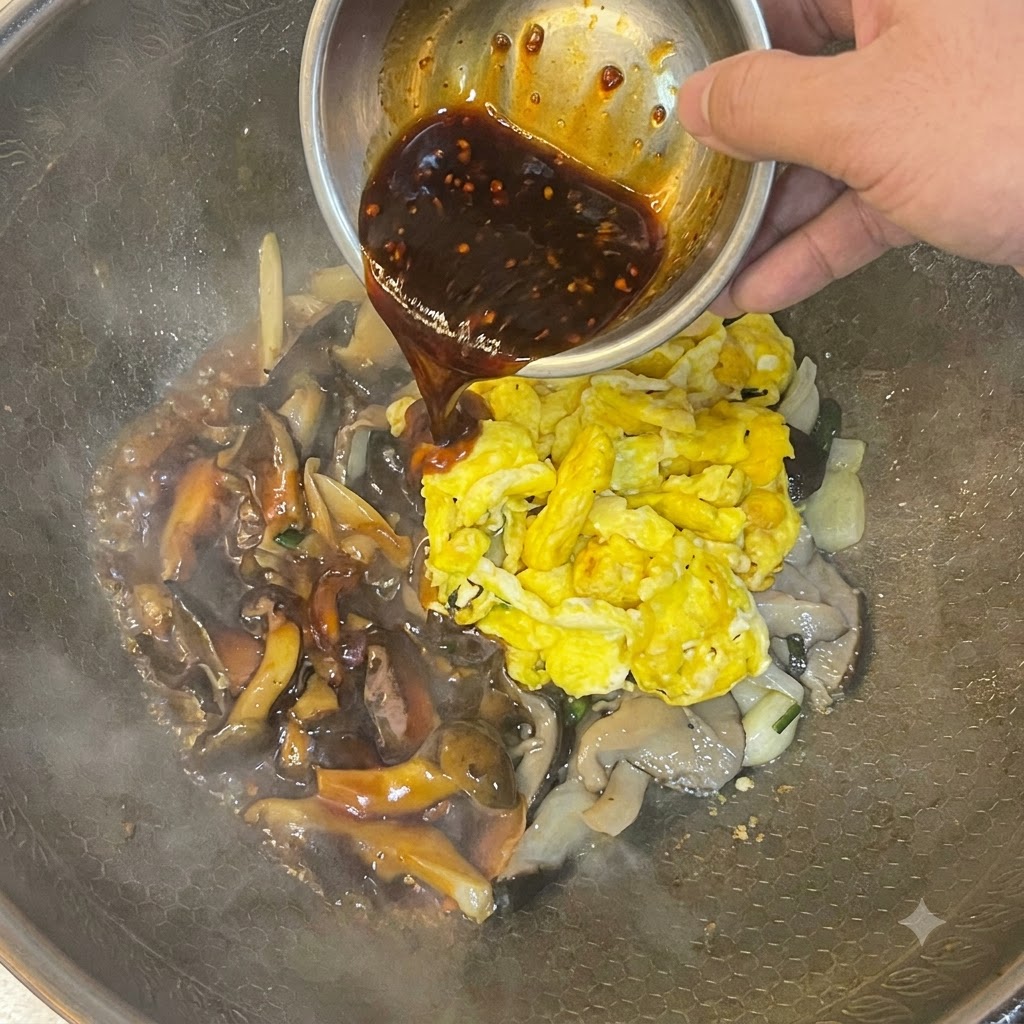 Dark sauce being poured from a metal bowl over scrambled eggs and mushrooms in a wok.