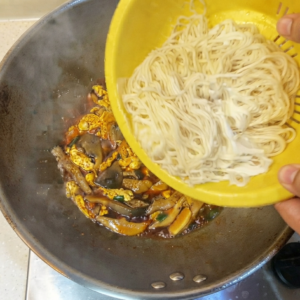 Cooked thin noodles being tipped from a yellow strainer into the wok containing sauce and ingredients.