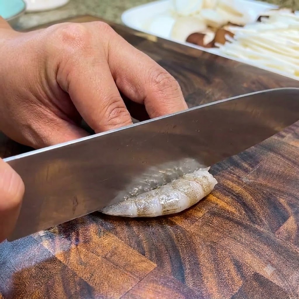 Raw shrimp being sliced and deveined with a knife on a wooden cutting board.