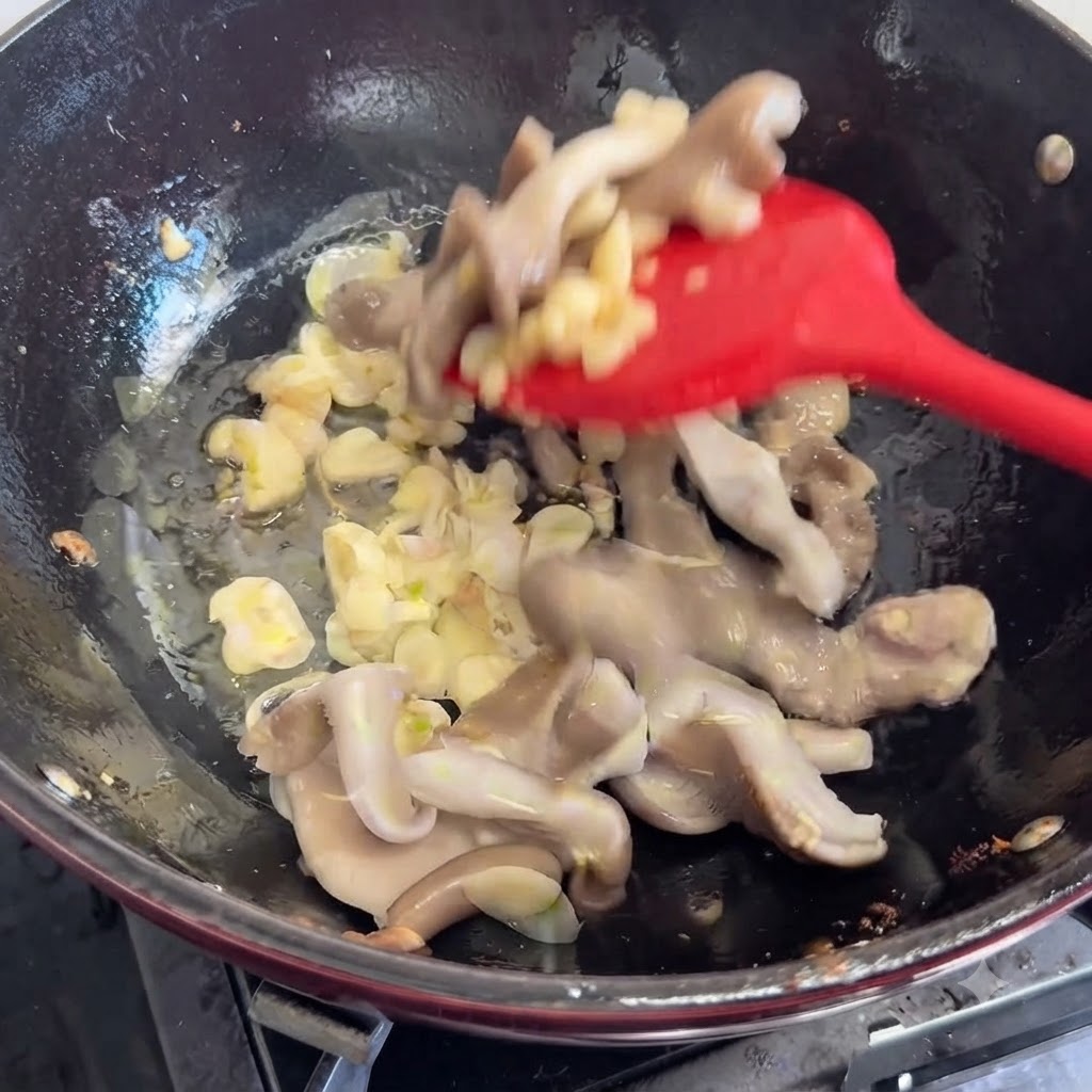 Fresh mushrooms being added and stirred in the pan with aromatics using a red spatula.