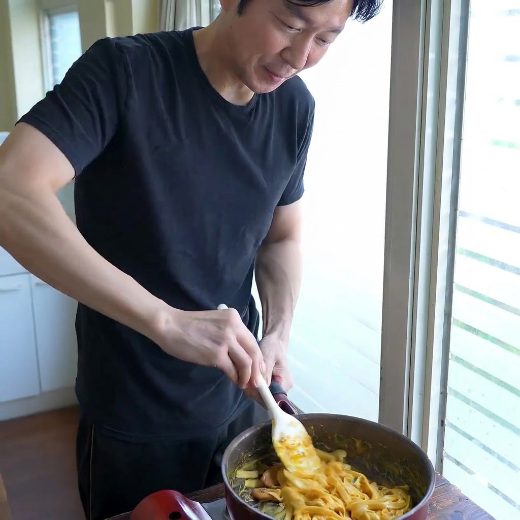 A person stirring wide noodles in a pan with a white spatula while adjusting the sauce consistency.