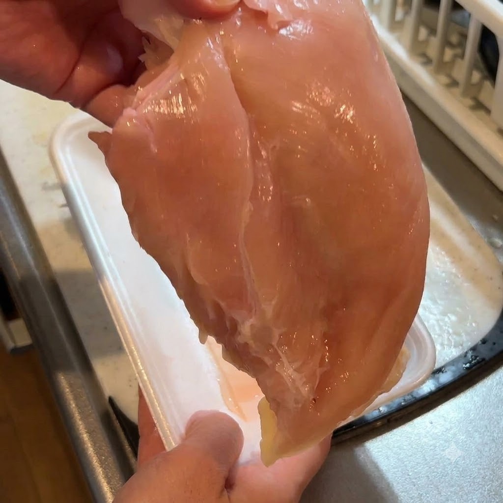 A raw chicken breast being held up while the skin is being removed with a knife.