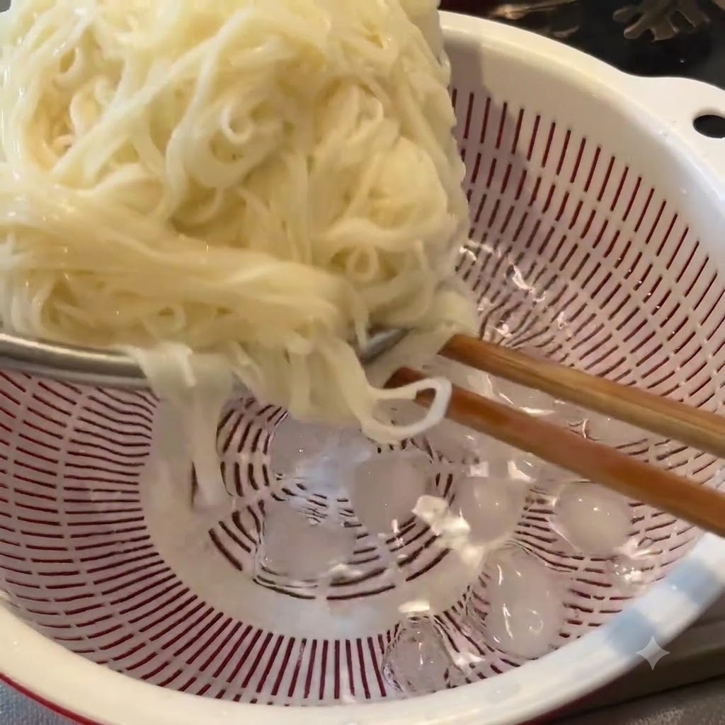 Cooked noodles being rinsed with chopsticks inside a white colander filled with ice cubes and cold water.
