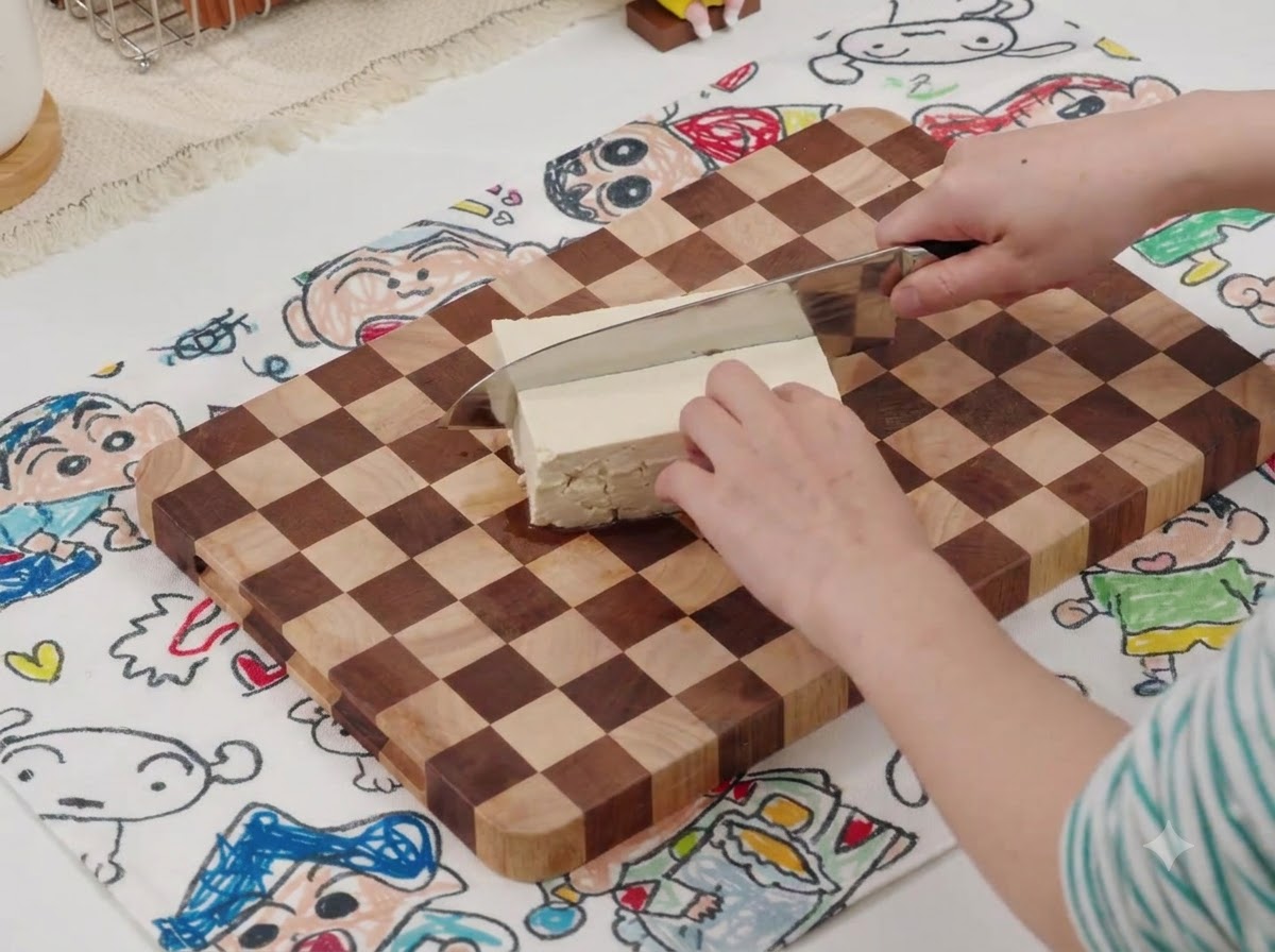 A knife slicing a block of white tofu on a wooden cutting board with a patterned mat underneath.