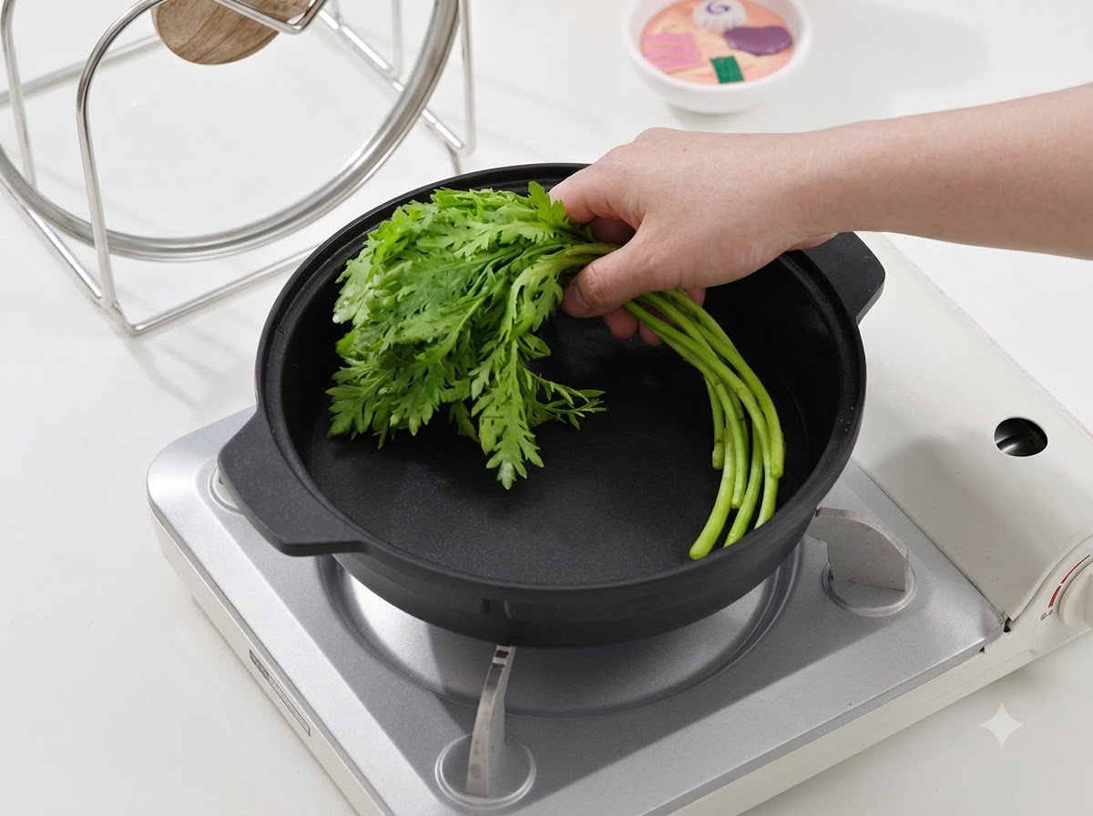 A hand placing a bundle of fresh green leafy vegetables into a black cooking pan on a portable stove.