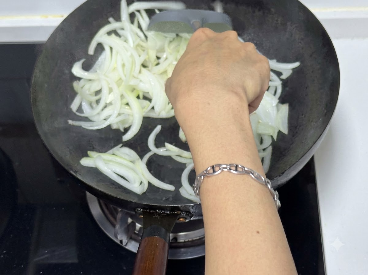 A person using a spatula to stir-fry sliced onions in a wok, showing translucent and slightly browned pieces.